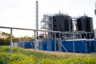 Wastewater Plant at DSM Industrial facility with several large black tanks and blue containers, surrounded by flowering yellow plants, under a cloudy sky, in connection with MBBR water treatment technology.