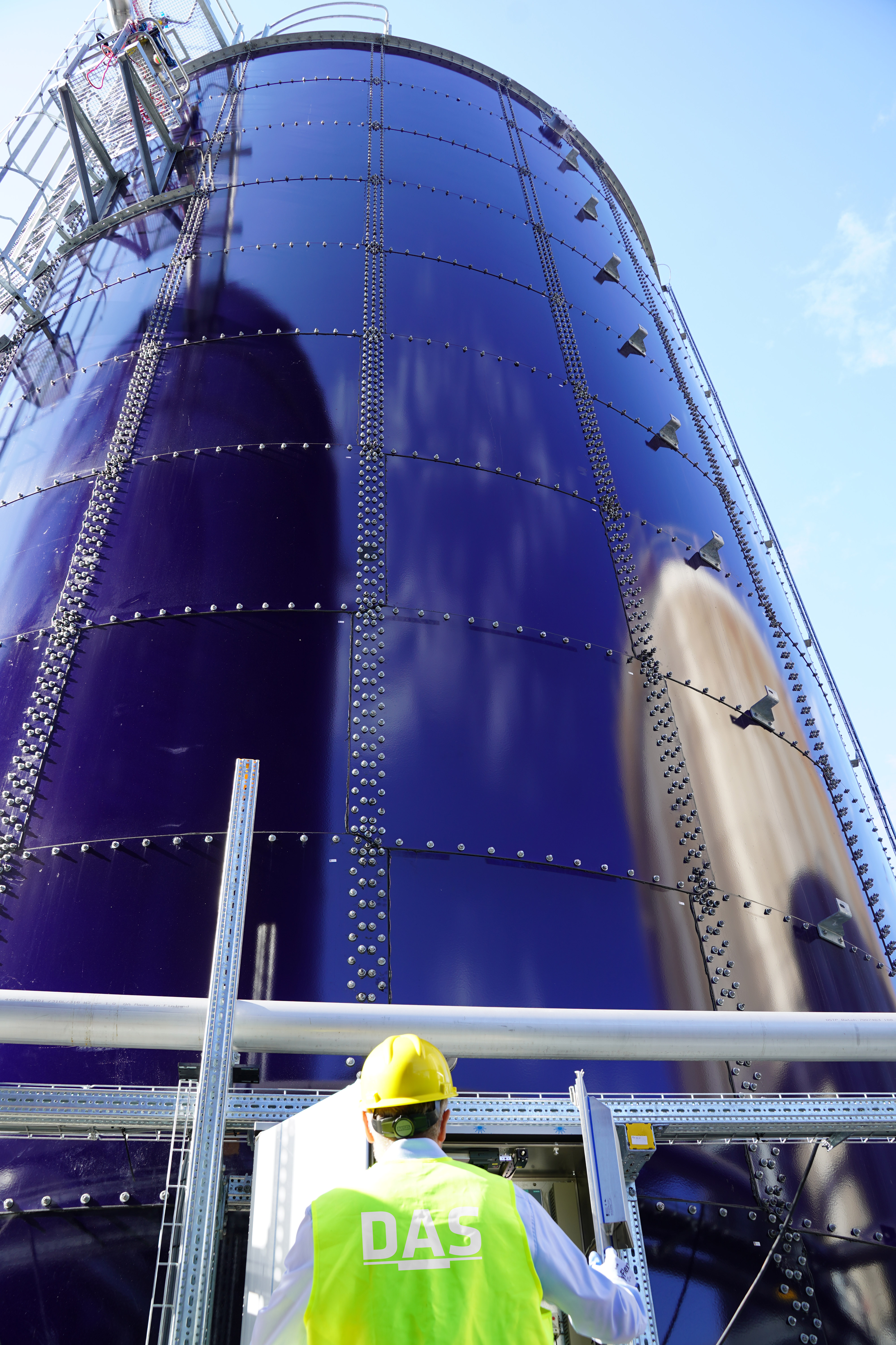 Ein Arbeiter von DAS EE mit Schutzhelm und reflektierender Weste steht vor einem hohen, modernen Silotank (MBBR-Anlage) aus glänzendem Metall unter klarem, blauem Himmel.