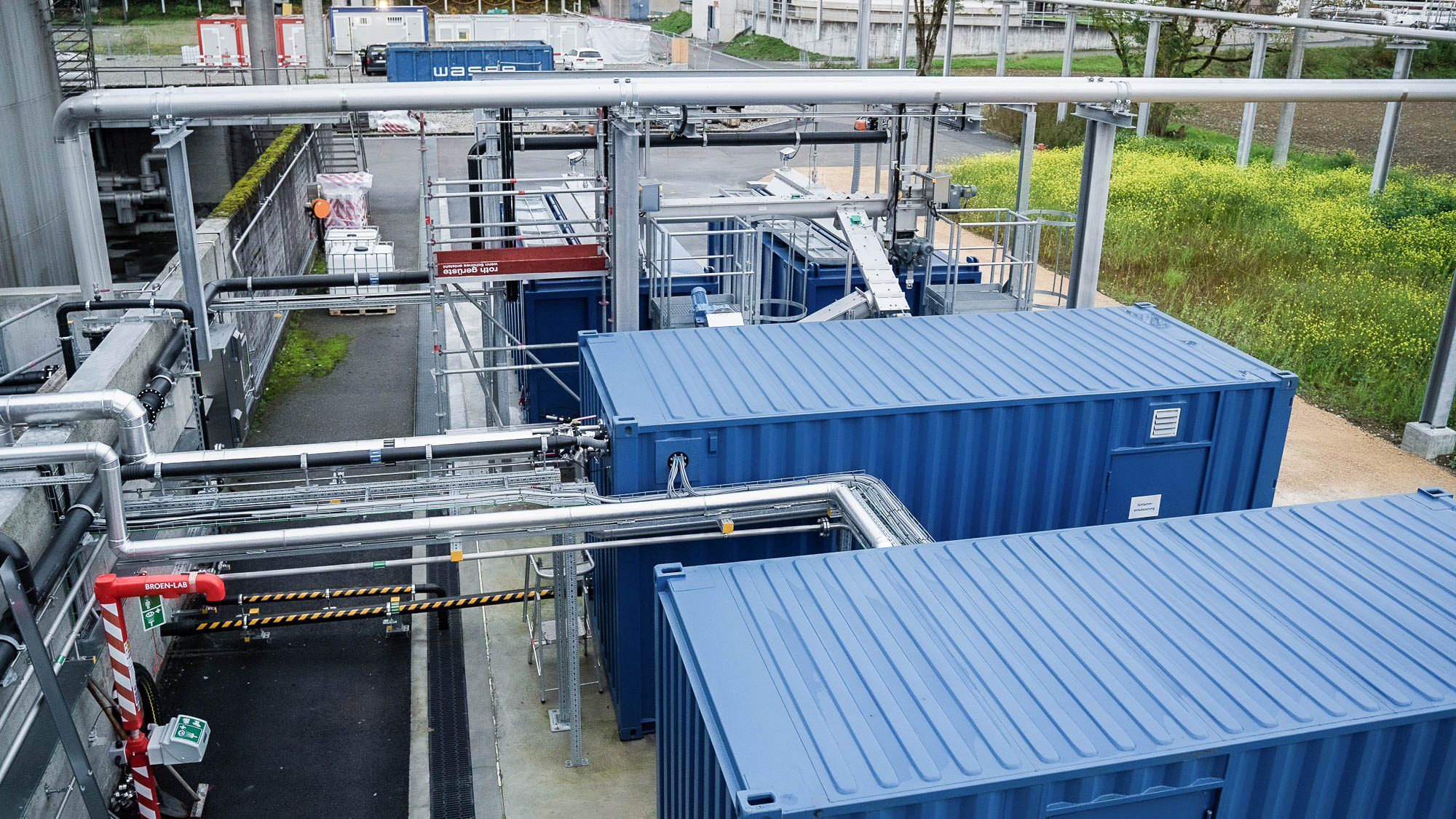 Blauer Container und industrielle Rohrinstallationen auf einem Betriebsgelände, umgeben von Pflanzen und grauen Gebäudestrukturen.