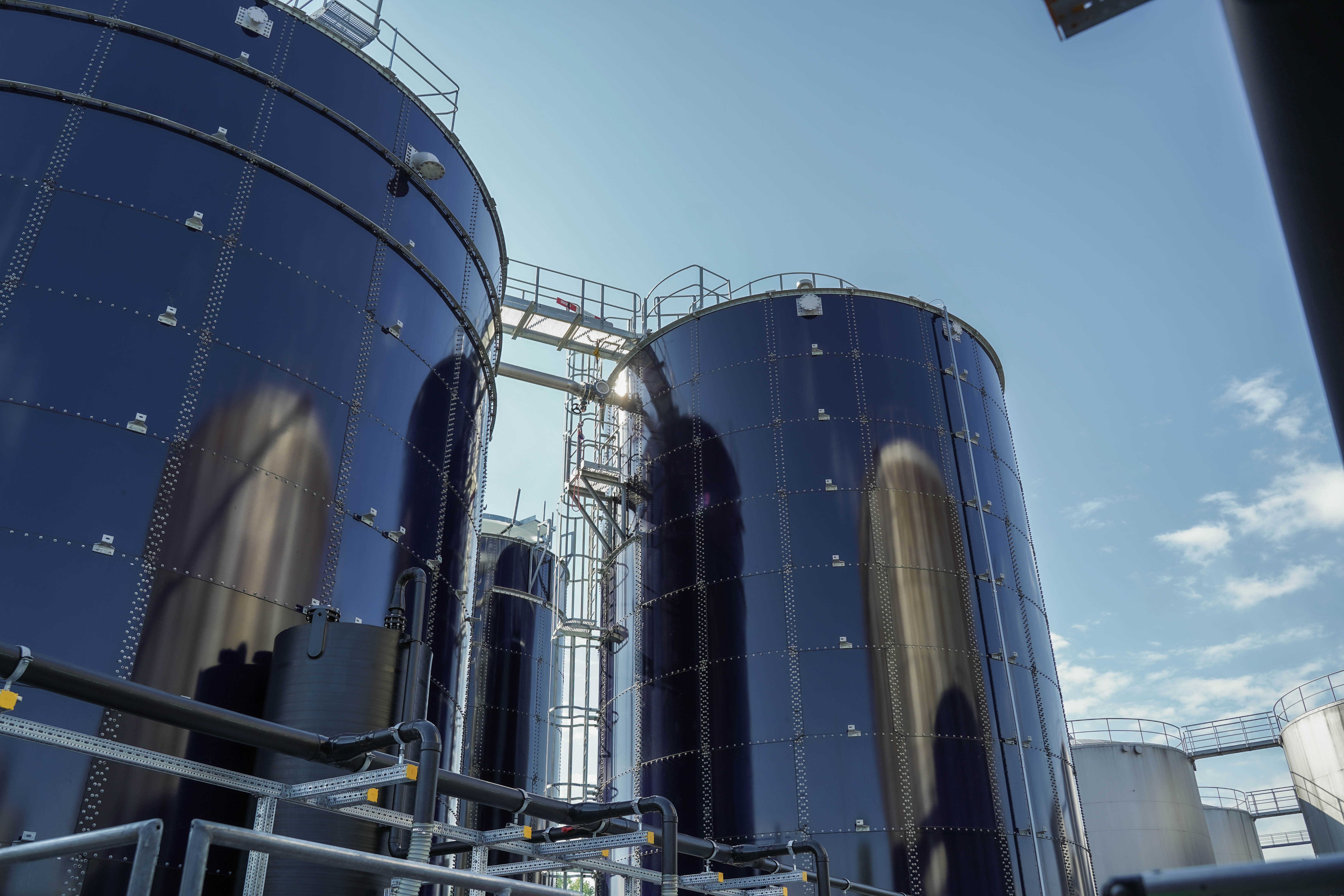 Große, zylindrische MBBR-Anlage-Tanks aus glänzendem Metall unter klarem, blauem Himmel, umgeben von industriellen Rohren und Leitungen auf einem modernen Abwasserbehandlungswerkgelände.