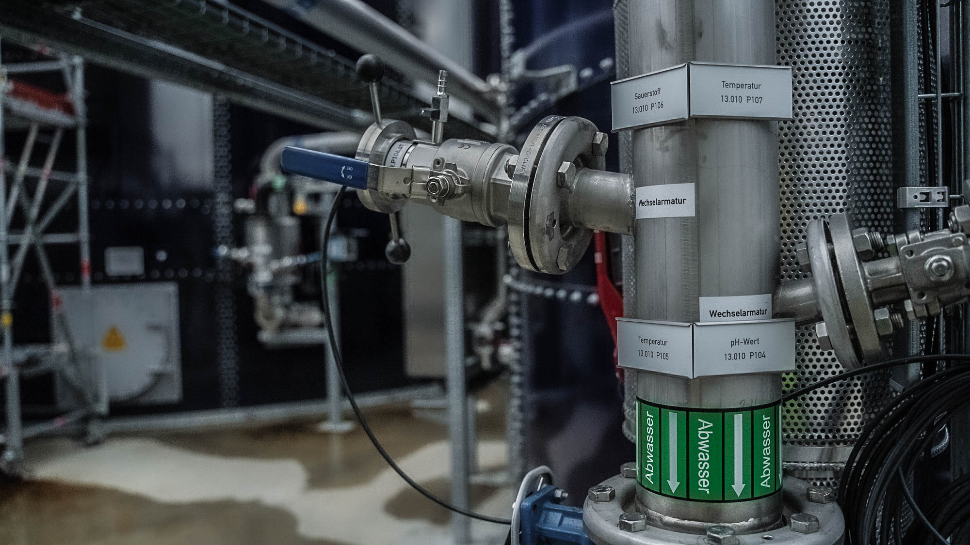 Industrielle Rohrleitung mit mehreren Ventilen und Schildern, auf denen Informationen wie "Standard", "Rücklauf im Filter" und "Abwasser" in einer technologischen Umgebung aufgedruckt sind.