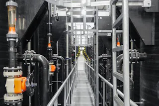 Kesslergrube Cover An industrial plant room with multiple pipes and valves flanks a metal maintenance walkway that runs through a modern factory environment.