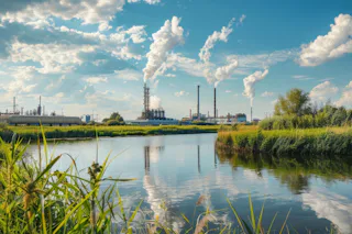Waste Gas Treatment An industrial landscape with several chimneys emitting smoke into the partly cloudy sky, while a tranquil river reflects the surroundings and blue sky, framed by green vegetation.