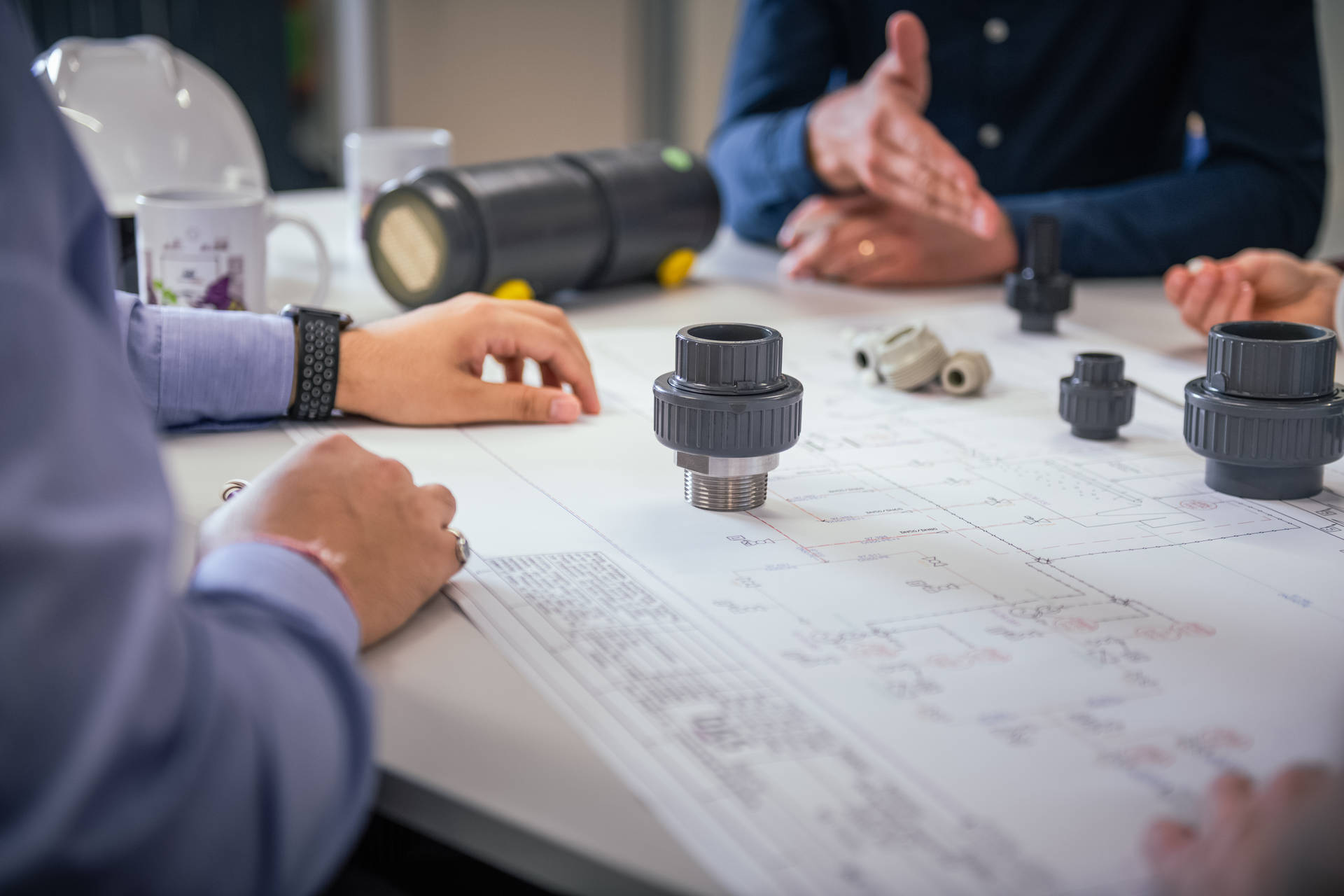 A group of people are discussing technical plans at a table on which various pipe fittings are laid out.