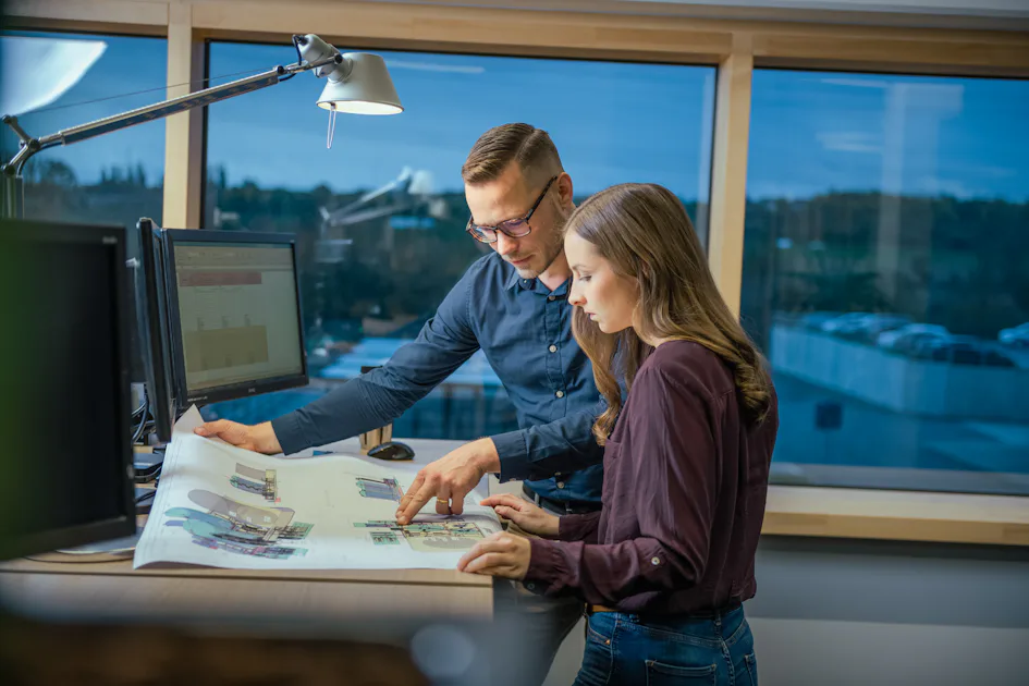 Precise planning: Our engineers develop forward-looking solutions. A man and a woman collaborate at a standing desk, examining architectural blueprints, surrounded by computer monitors in a modern office setting with large windows providing a view of the outdoors.