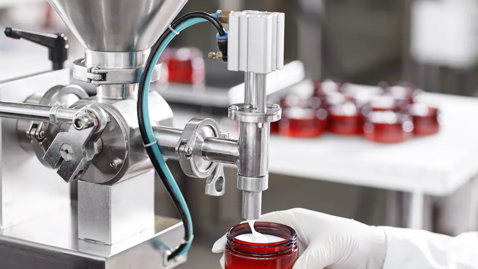 A worker in protective gloves operates a machine filling red jars with a creamy substance in a cosmetics production facility, surrounded by other filled jars on a white table.
