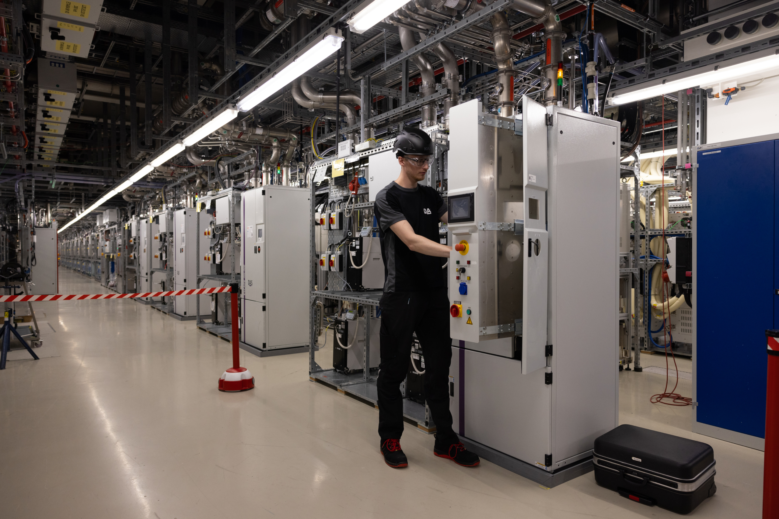 A technician wearing safety goggles works on a control cabinet in a modern, well-lit subfab, surrounded by a long row of technical equipment and cable harnesses.