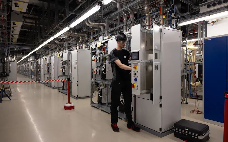 Technician standing at a burn-wet system A technician wearing safety goggles works on a control cabinet in a modern, well-lit subfab, surrounded by a long row of technical equipment and cable harnesses.