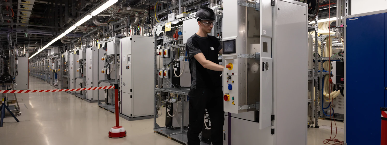 Technician standing at a burn-wet system A technician wearing safety goggles works on a control cabinet in a modern, well-lit subfab, surrounded by a long row of technical equipment and cable harnesses.