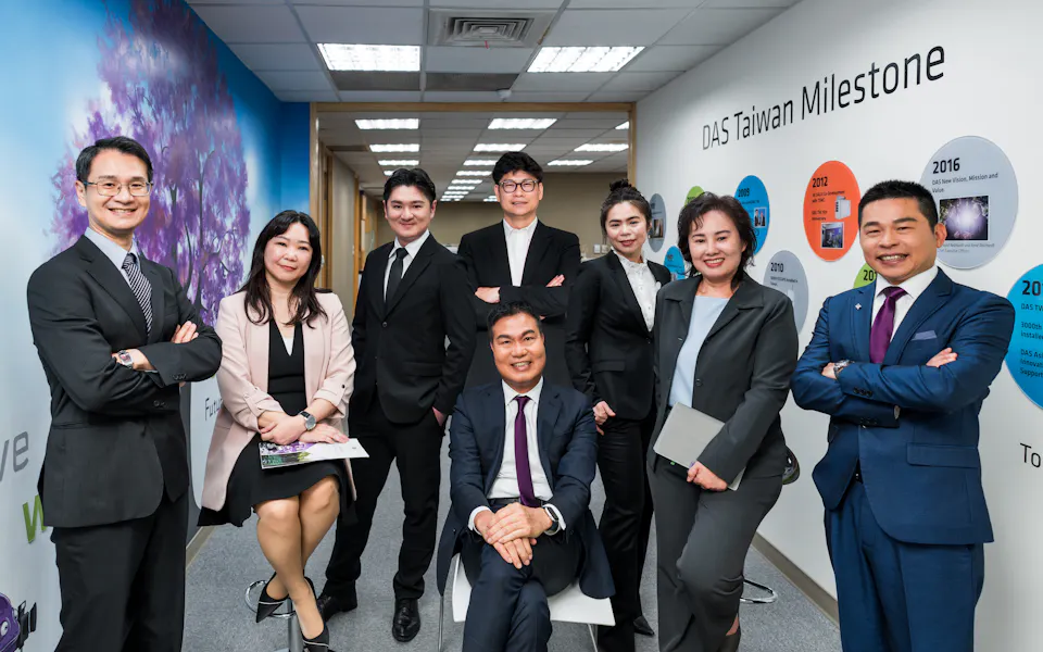 management team 這張圖片顯示了一群穿著正式商務服裝的專業人士在一間現代化辦公室合影,背景是一面牆,上面展示了多個代表台灣里程碑的圓形資訊圖表。