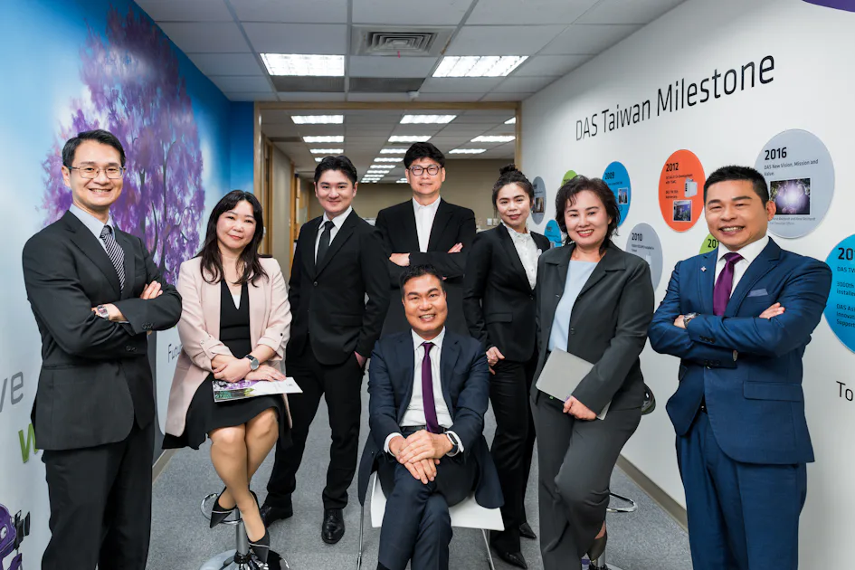 management team 這張圖片顯示了一群穿著正式商務服裝的專業人士在一間現代化辦公室合影,背景是一面牆,上面展示了多個代表台灣里程碑的圓形資訊圖表。
