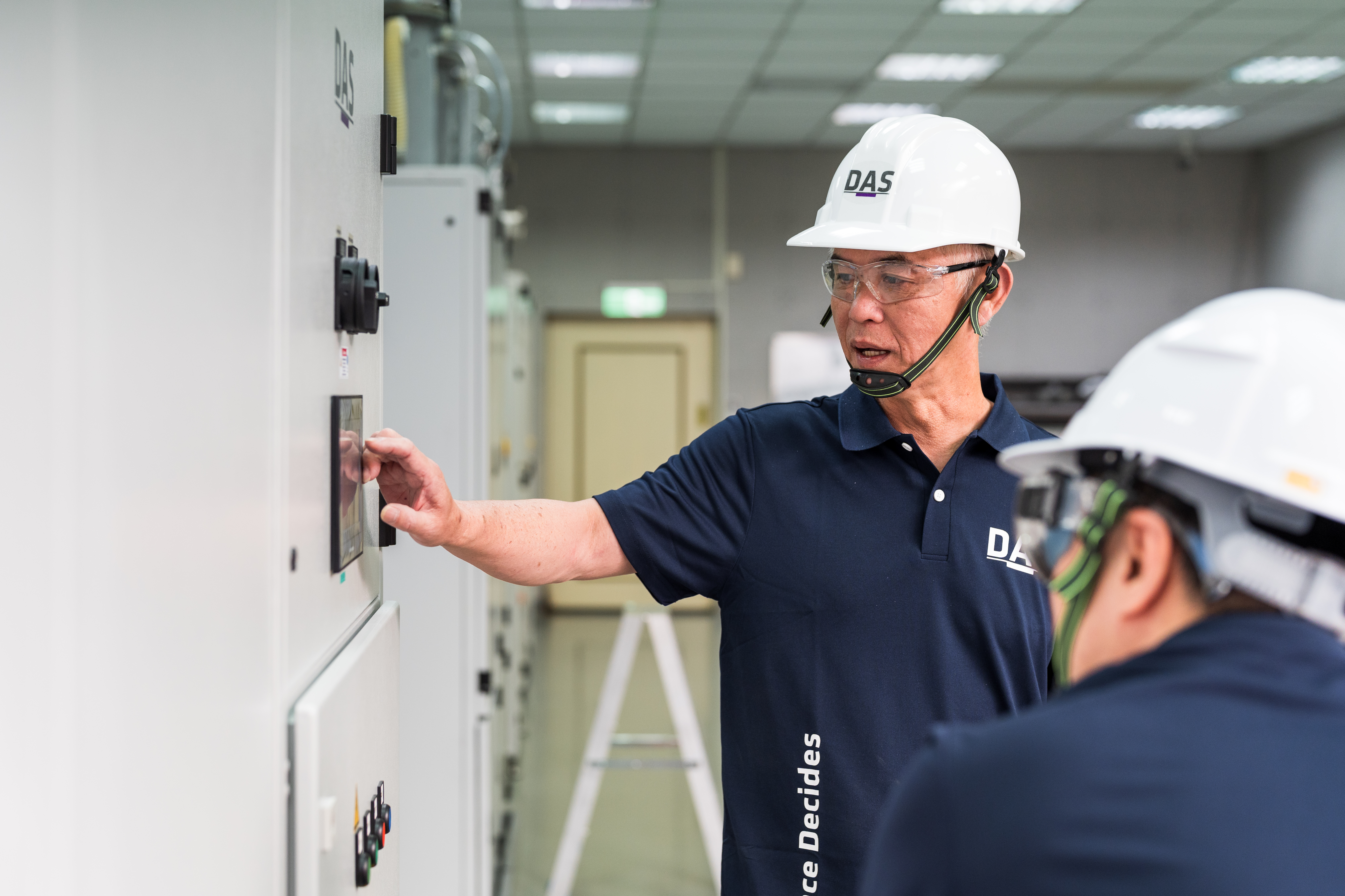 兩名穿著深藍色制服和安全帽的工人在工廠內操作大型電控面板，一人正在觸控面板並查看其顯示內容，周圍環境整潔且照明明亮。