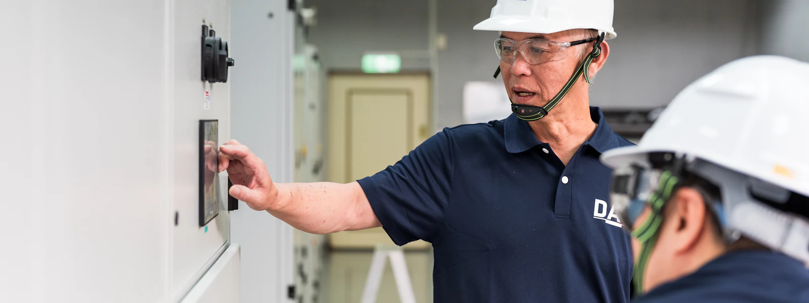 ESCAPE 兩名穿著深藍色制服和安全帽的工人在工廠內操作大型電控面板,一人正在觸控面板並查看其顯示內容,周圍環境整潔且照明明亮。