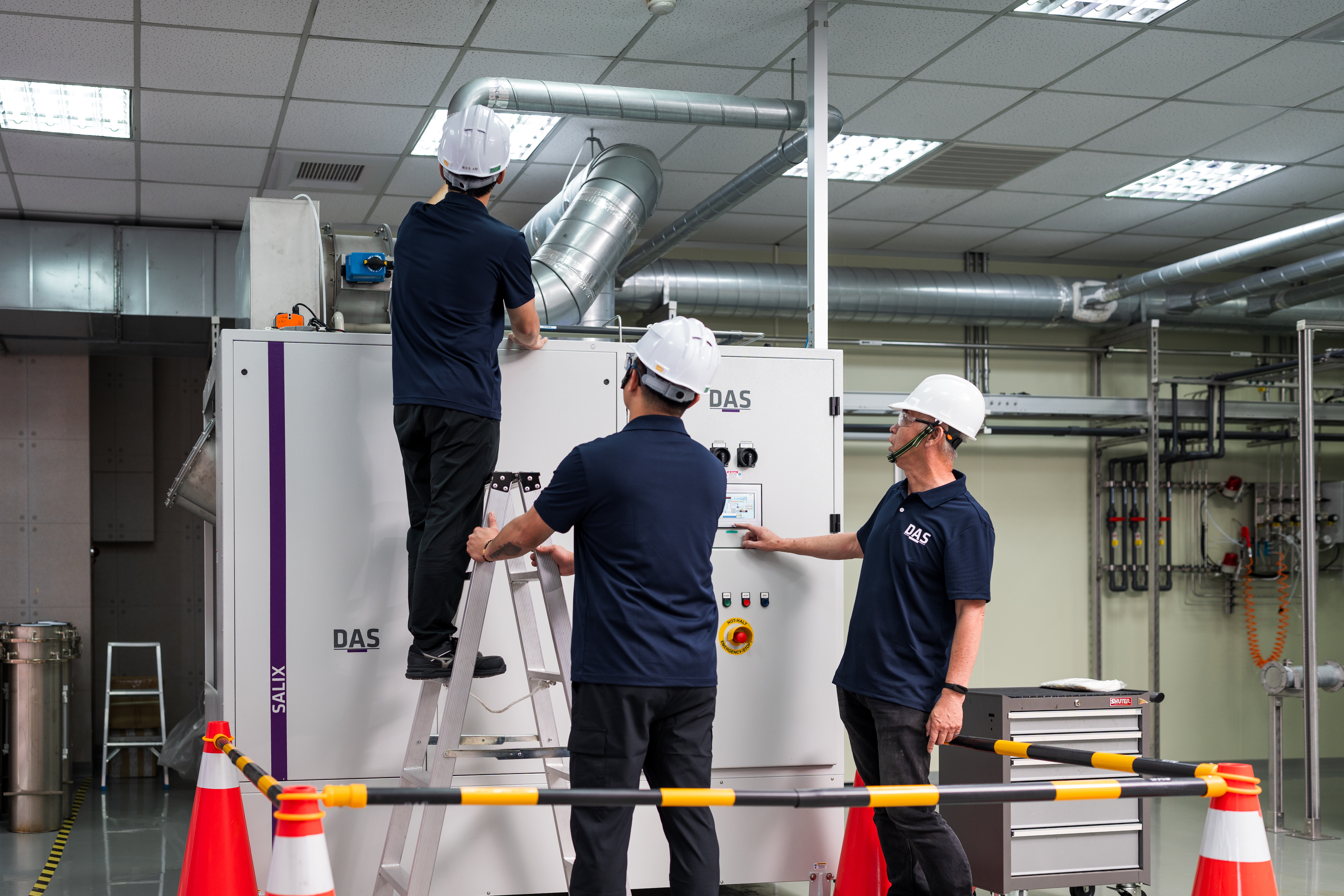 三位戴著安全帽的人員在明亮的工業設施中，操作大型金屬設備，專注於機器的檢查和維護，周圍有橙白相間的安全欄杆。