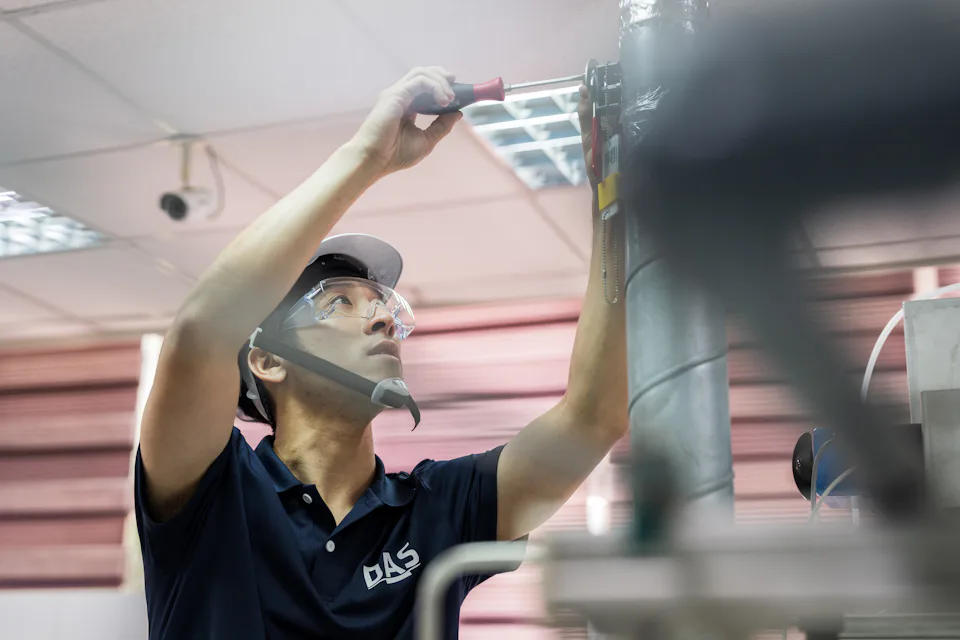 Ein DAS-Techniker mit Schutzbrille und Helm arbeitet mit einem Schraubendreher an einem Abgasreinigungssystem in einer industriellen Umgebung.