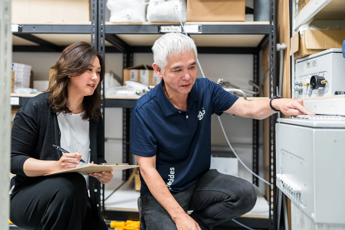 Mitarbeiter Taiwan Two people are collaborating in a storage area, with one person taking notes on a clipboard while the other points to equipment on a shelf, surrounded by boxes and technical devices.