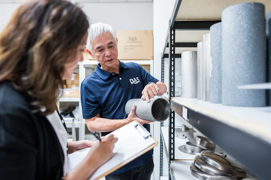 parts 一位短髮銀髮男性正在貨架前向一位持筆記板的長髮女性介紹管道,背景是工業倉庫,貨架上擺滿各種管道產品。