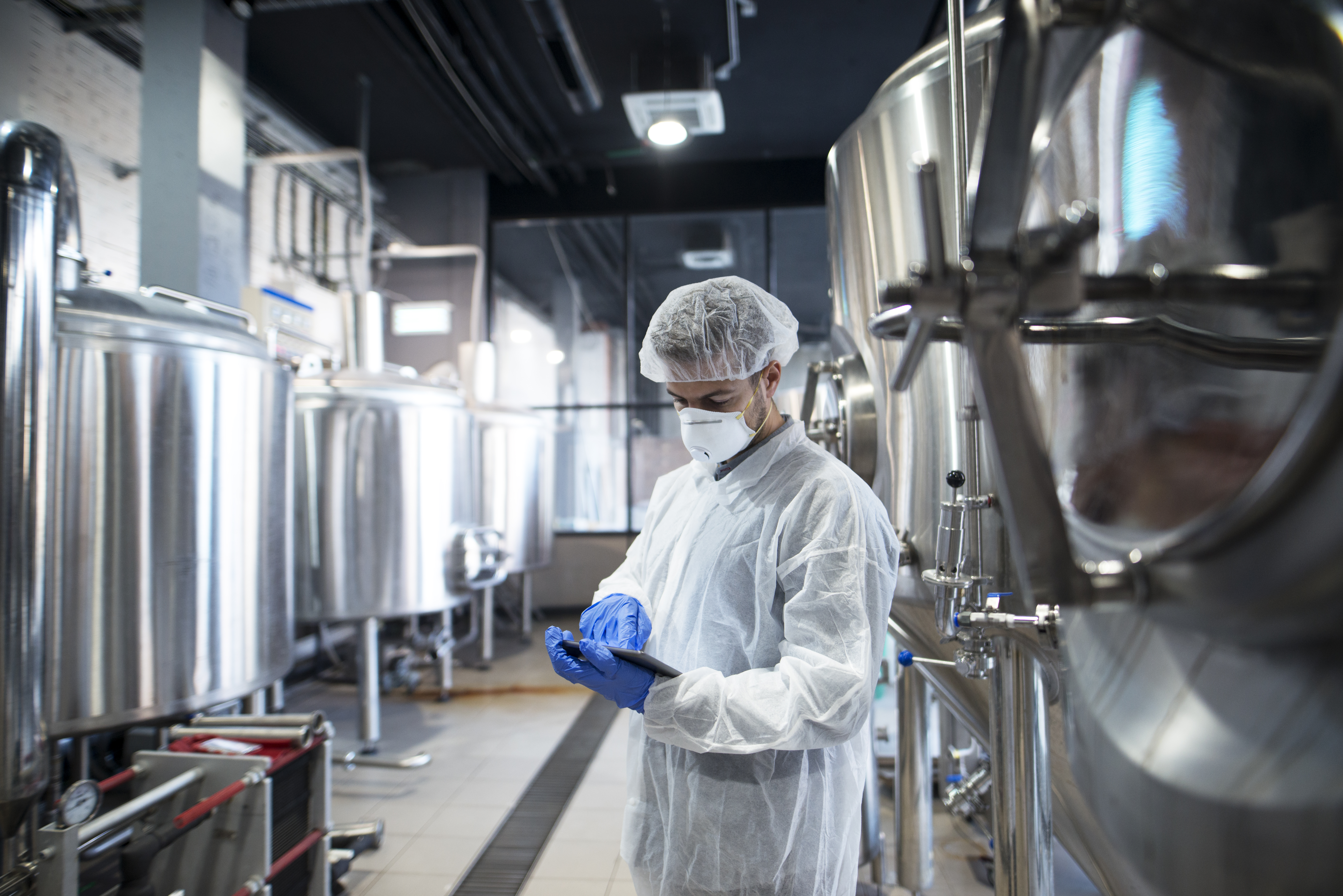 A worker in a white protective suit and mask inspects equipment in a modern brewery with large stainless steel fermentation tanks, highlighting brewery operations and safety standards.