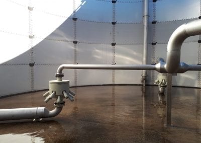 A large, round industrial tank (MBBR) with visible galvanized steel pipes and an ejector on a wet concrete floor inside.