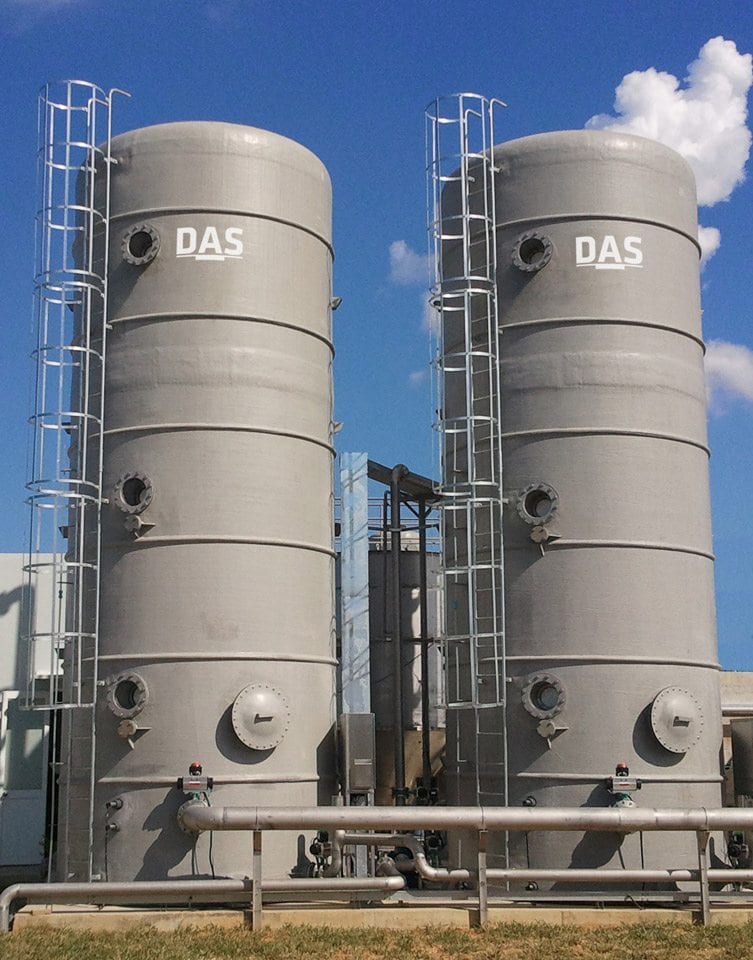 The image shows two large industrial grey storage tanks labeled with "DAS," standing side by side under a clear blue sky, connected by a network of metallic pipes and surrounded by a grassy area.