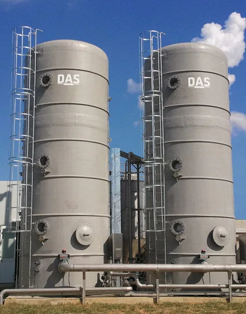 Käseherstellung The image shows two large industrial grey storage tanks labeled with "DAS," standing side by side under a clear blue sky, connected by a network of metallic pipes and surrounded by a grassy area.