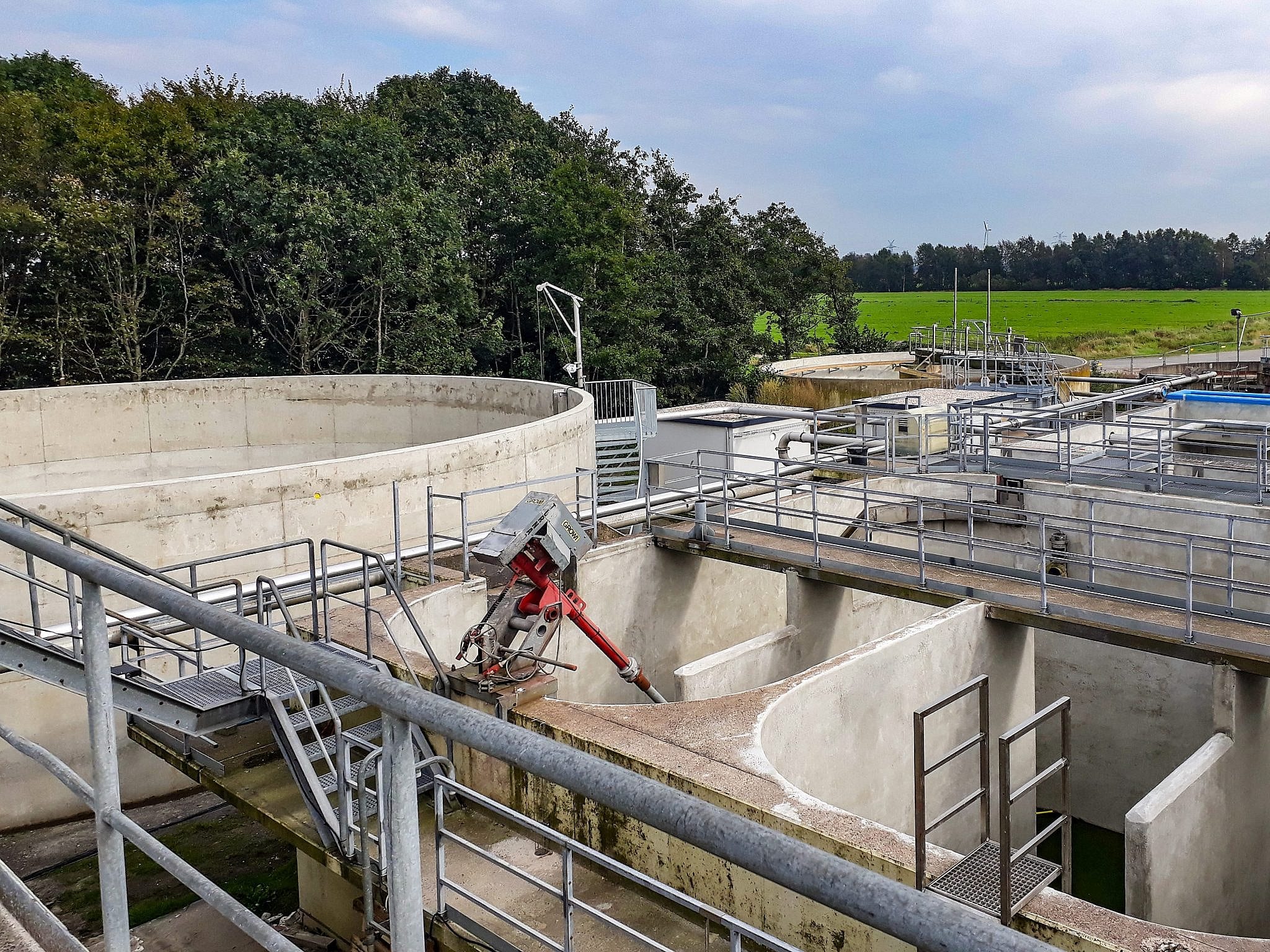 An industrial wastewater treatment plant featuring concrete tanks and metal walkways is surrounded by lush green fields and dense trees under a cloudy sky.
