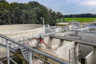Case Study DMK An industrial wastewater treatment plant featuring concrete tanks and metal walkways is surrounded by lush green fields and dense trees under a cloudy sky.