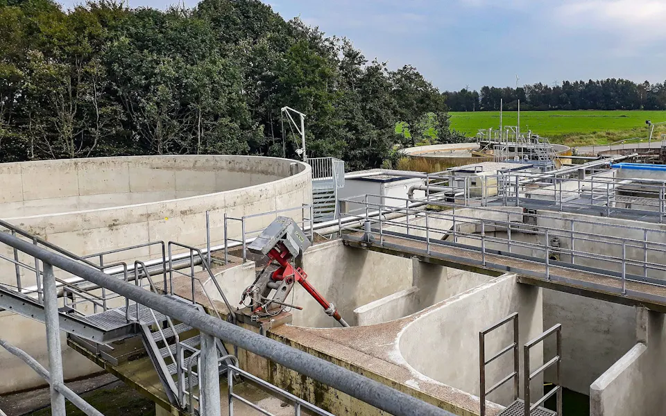 Case Study DMK An industrial wastewater treatment plant featuring concrete tanks and metal walkways is surrounded by lush green fields and dense trees under a cloudy sky.