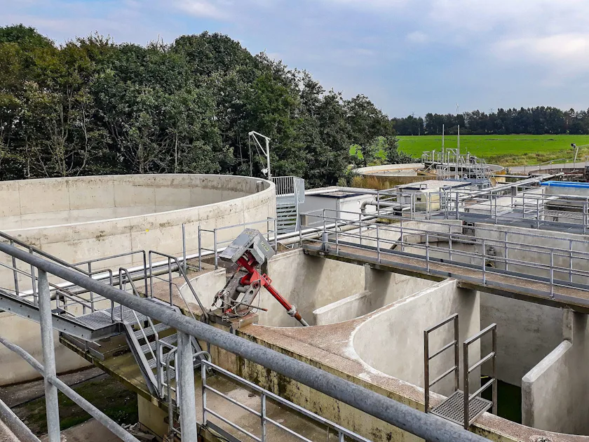 Fallstudie DMK An industrial wastewater treatment plant featuring concrete tanks and metal walkways is surrounded by lush green fields and dense trees under a cloudy sky.