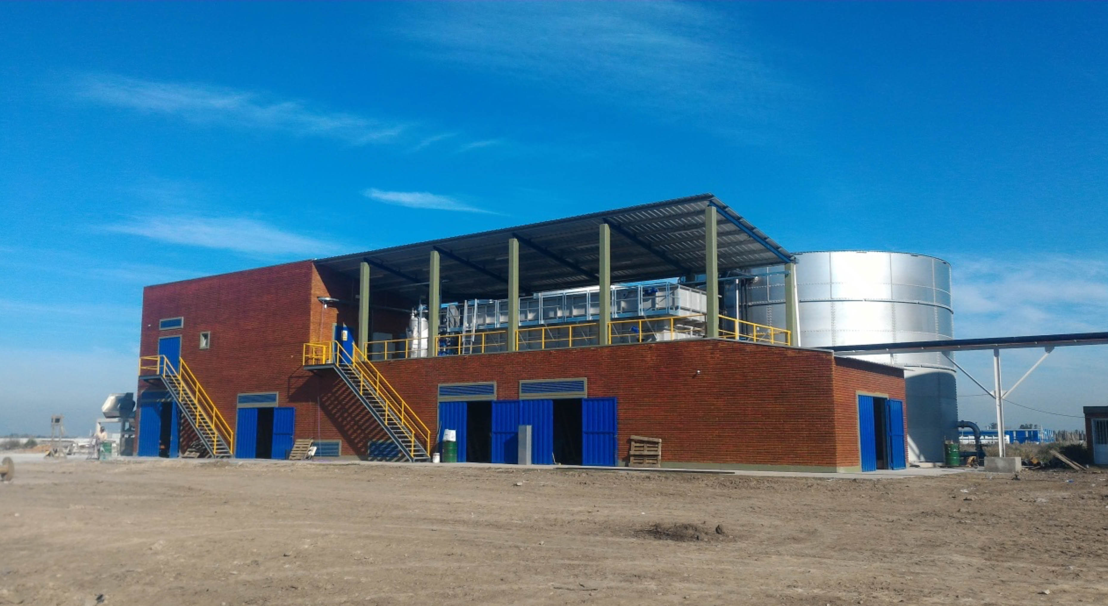 Building of a beverage manufacturer with red brick, blue doors and windows, and a silver water tank in the background under a clear blue sky.