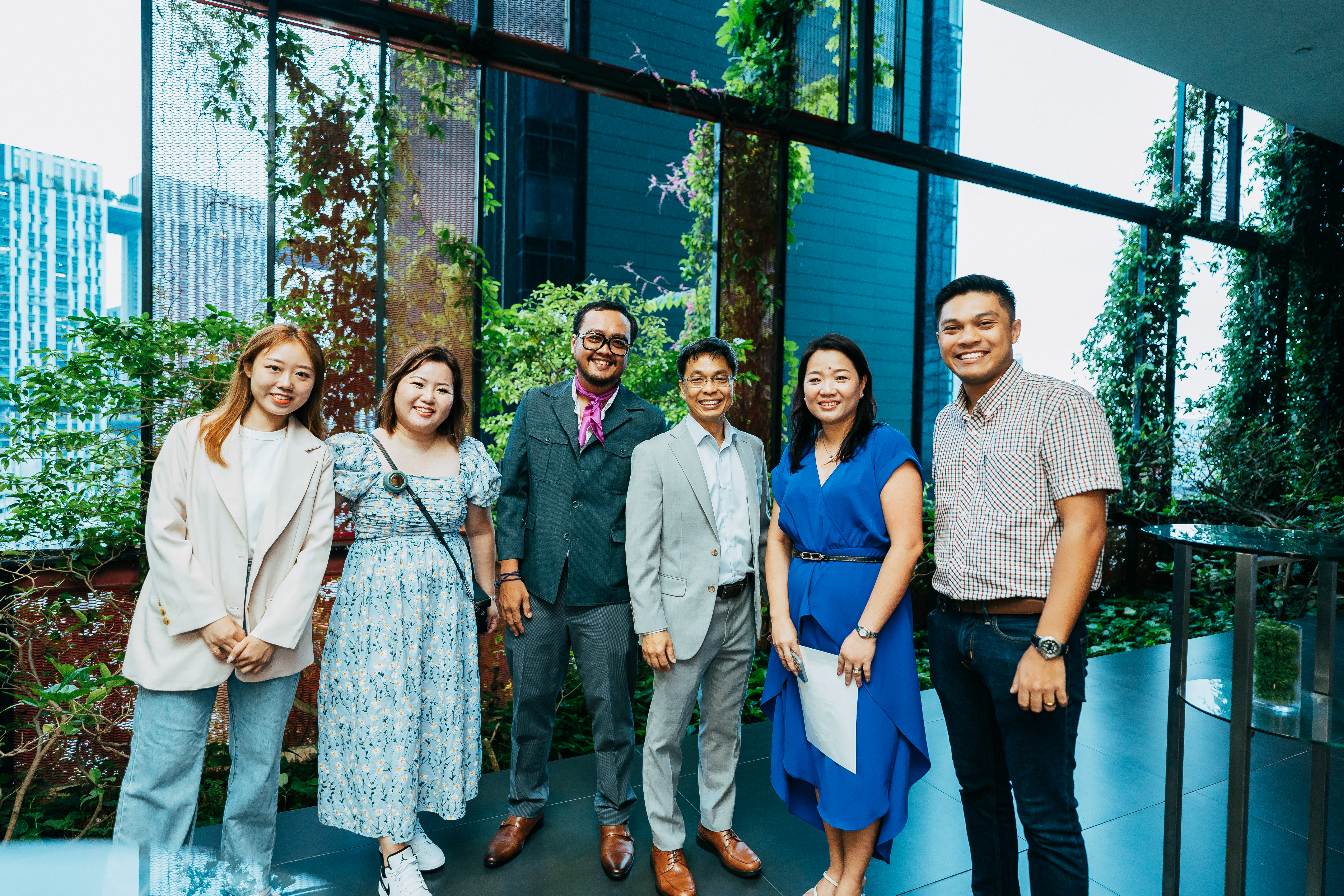 A diverse group of six people pose together, smiling warmly, in a modern, plant-filled office setting with large glass windows and a city view in the background.