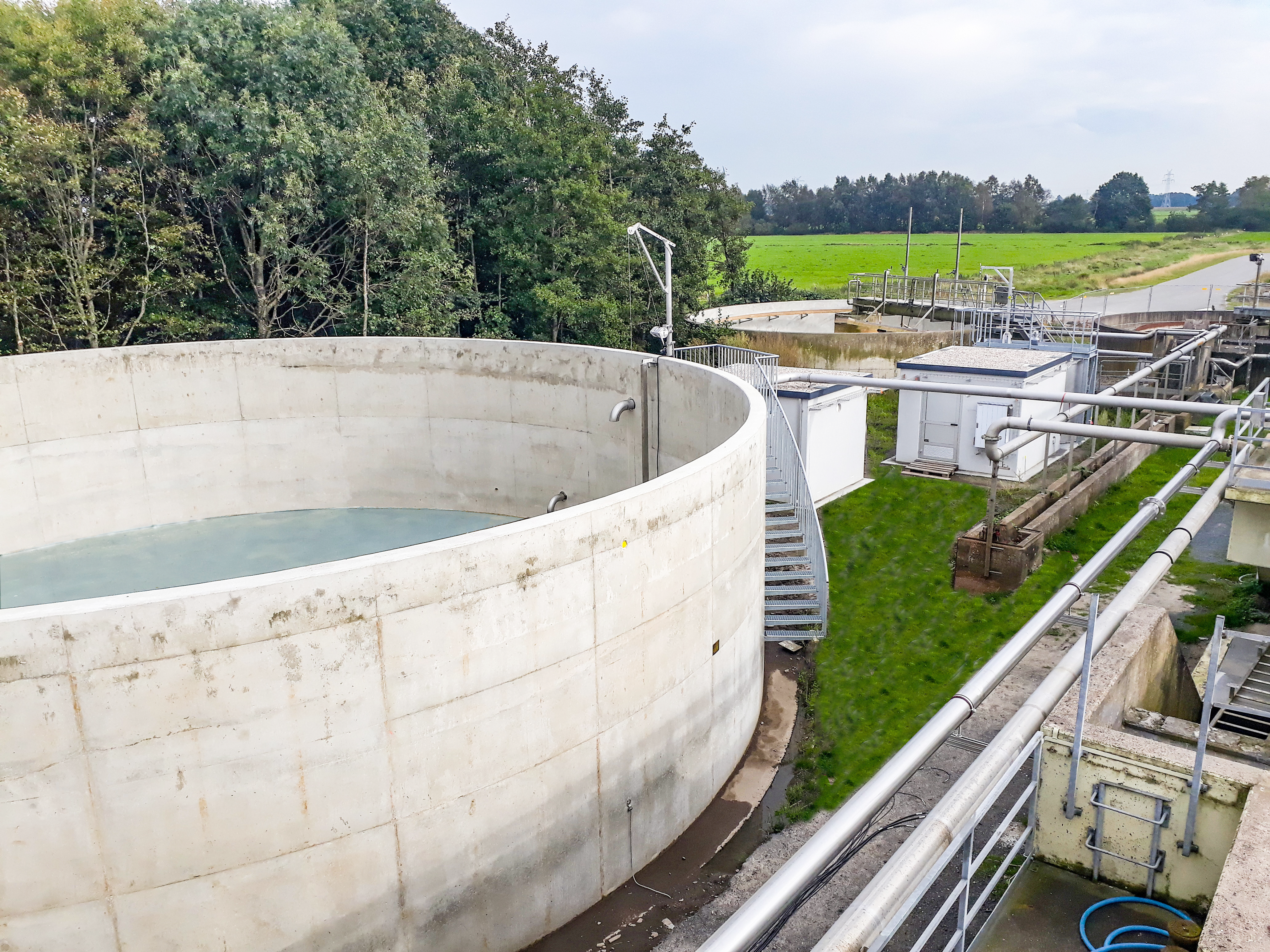 A large, round concrete water basin in a sewage treatment plant, surrounded by green fields and trees, with pipes and technical structures in the background.