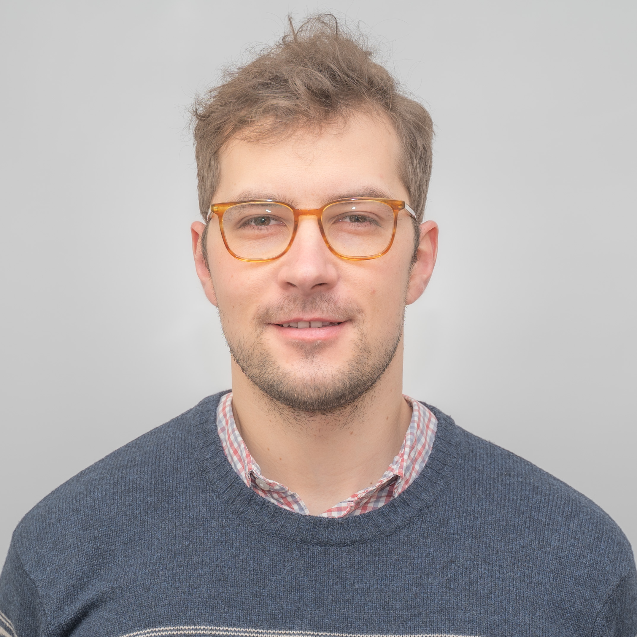 A person with short hair and glasses is wearing a blue sweater over a plaid shirt, standing against a plain gray background, embodying a professional and approachable style.