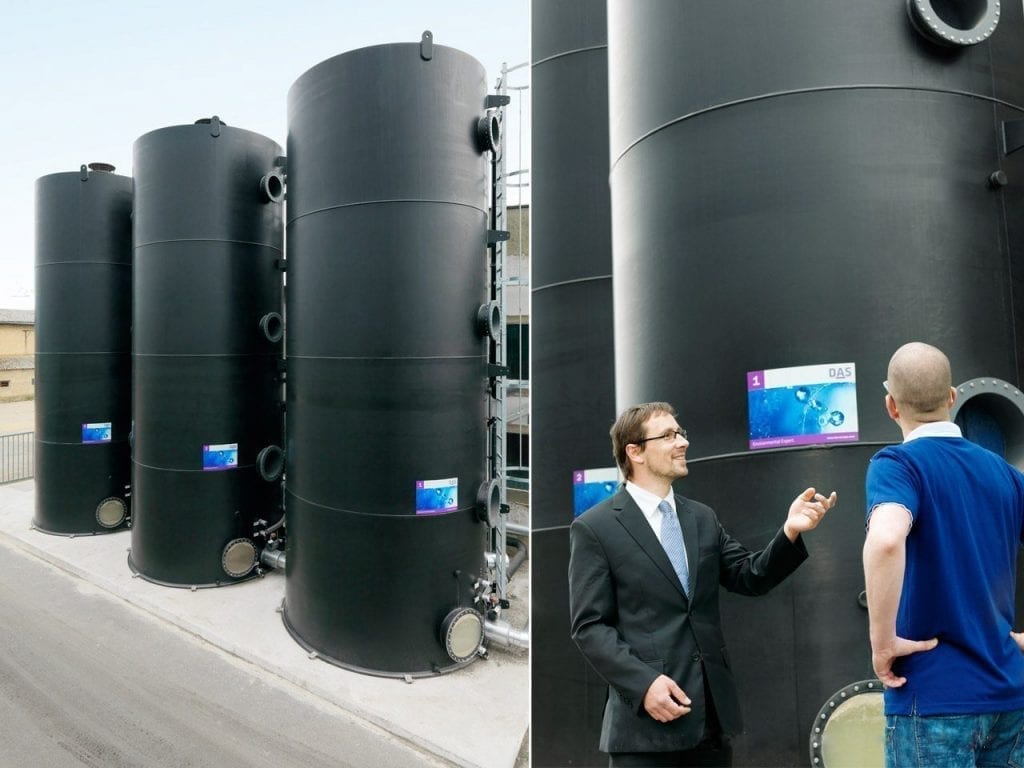 Three large black steel tanks stand side by side on a gray industrial site, while two men in formal attire stand in front of them talking to each other.