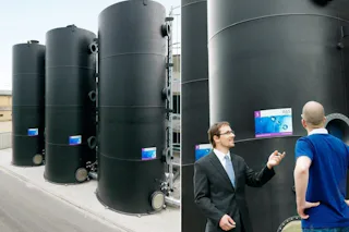 TFR Bioreactor Three large black steel tanks stand side by side on a gray industrial site, while two men in formal attire stand in front of them talking to each other.