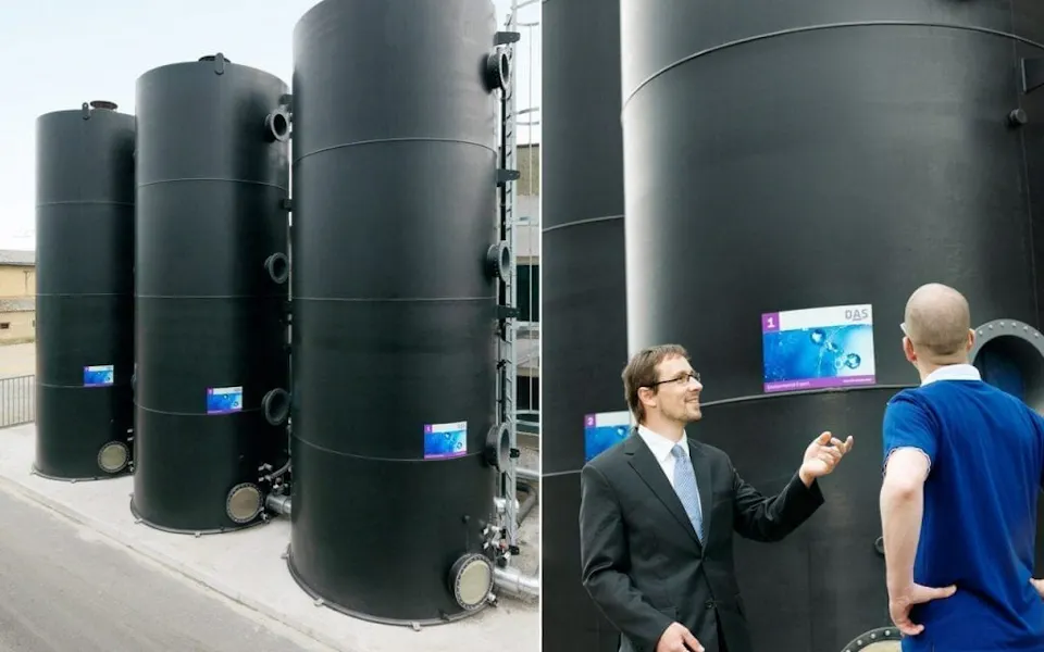 TFR Bioreactor Three large black steel tanks stand side by side on a gray industrial site, while two men in formal attire stand in front of them talking to each other.
