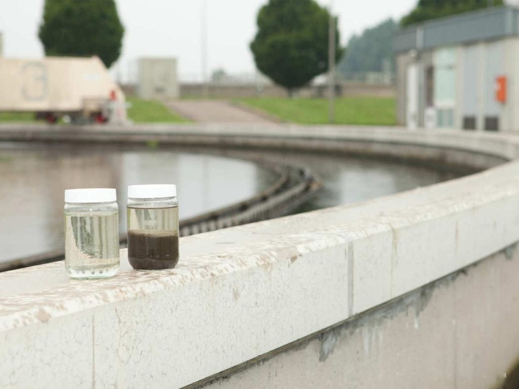 Two glass containers filled with water samples, one clear and the other cloudy, stand on a concrete balustrade in front of a sewage treatment plant basin.