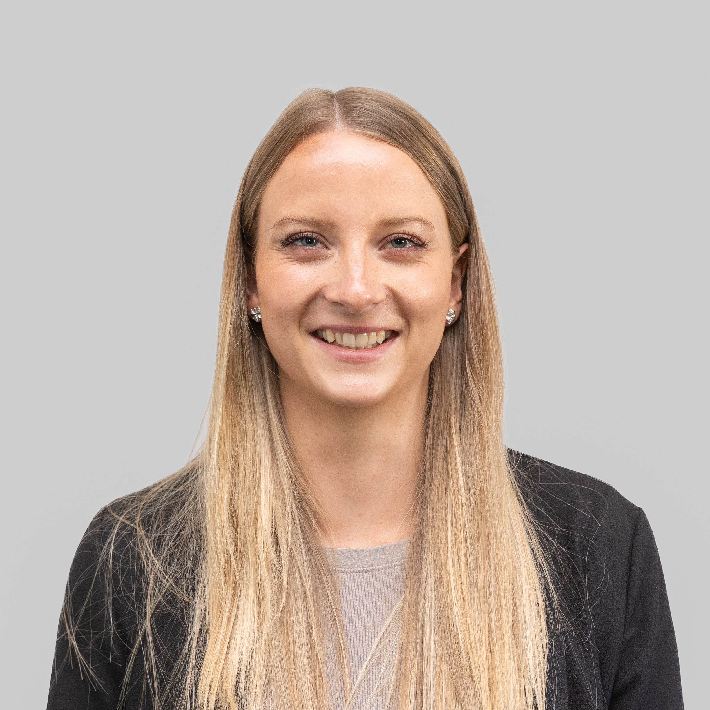 A professional woman with long blond hair is smiling confidently against a plain gray background, wearing a black blazer over a light-colored top, representing a technical project manager.