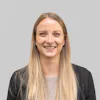 Carolin Siegert A professional woman with long blond hair is smiling confidently against a plain gray background, wearing a black blazer over a light-colored top, representing a technical project manager.