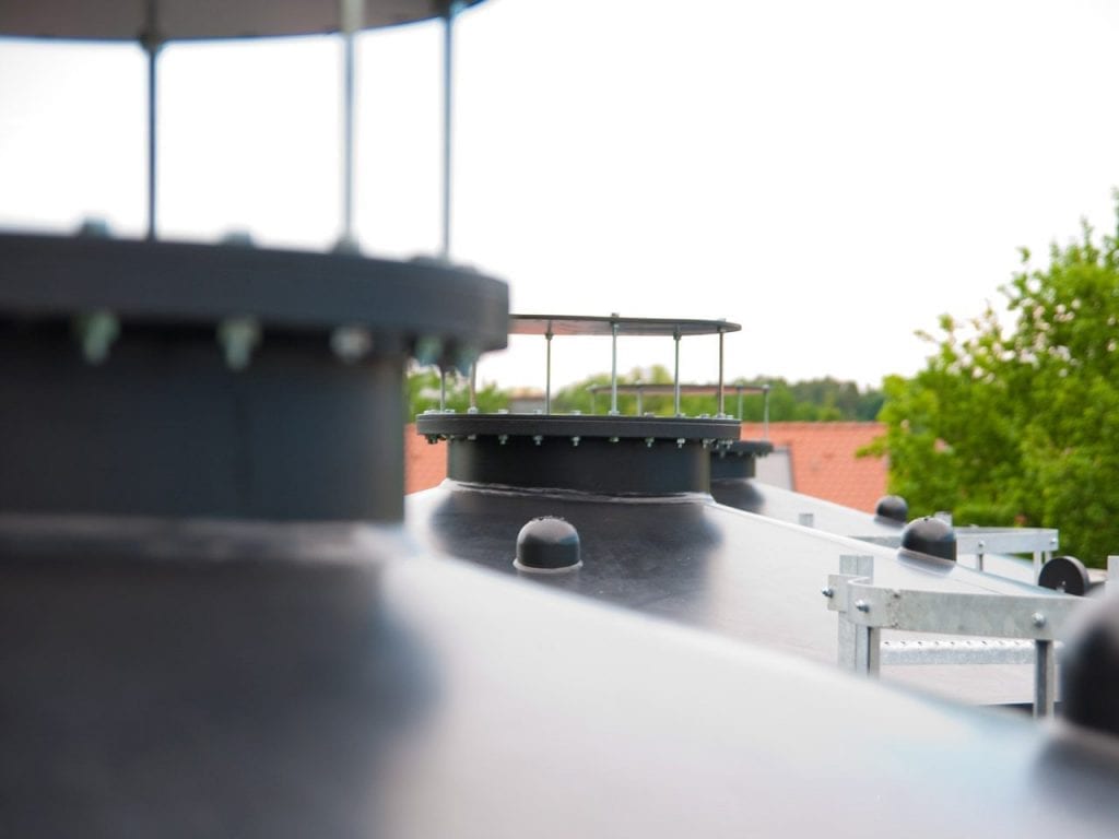 The image shows the top of large black tanks with metal caps, standing outdoors in an industrial setting, surrounded by trees and a brick building in the background.