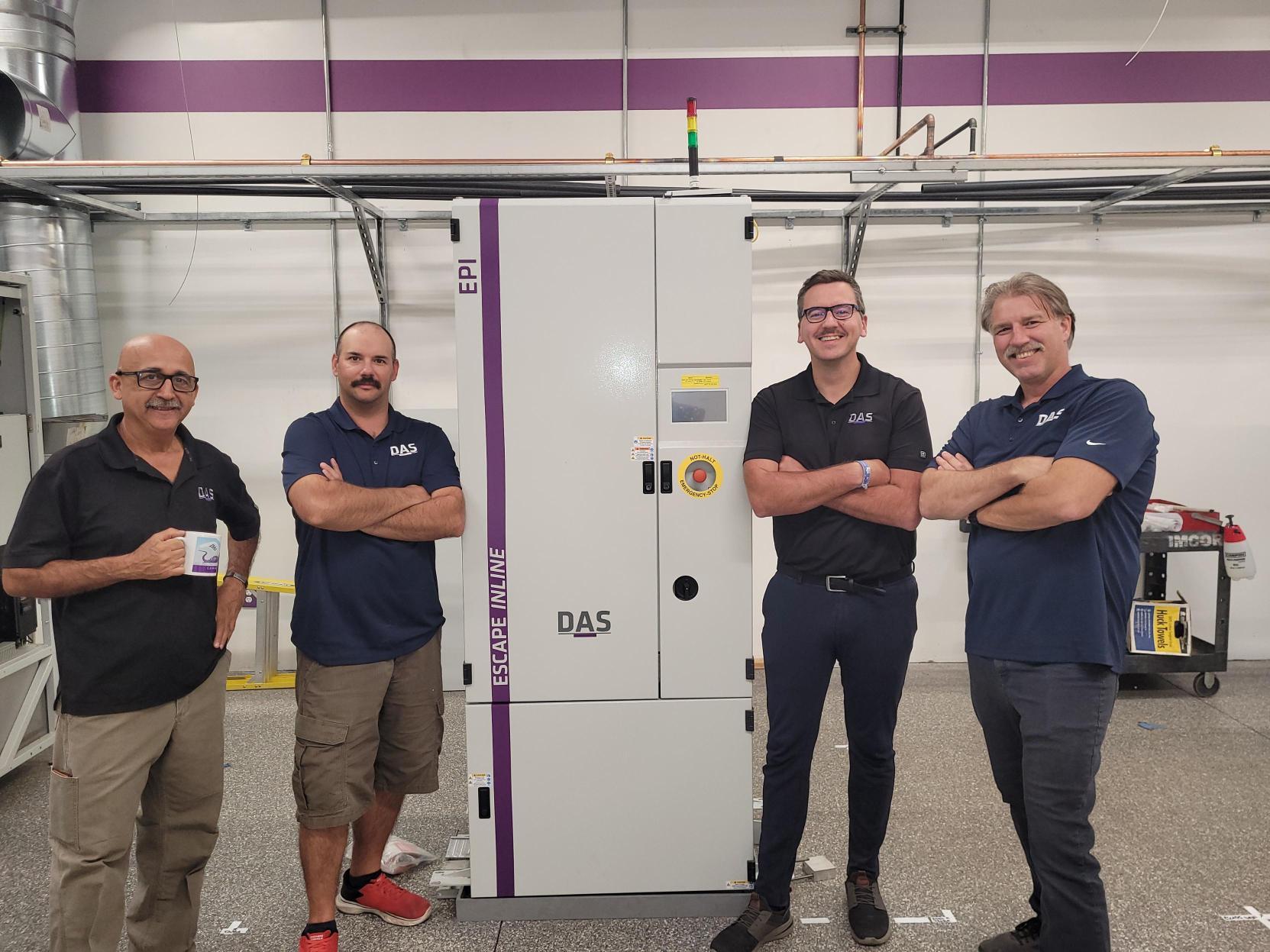 Four men in matching navy shirts stand confidently in front of a large industrial control panel, labeled "DAS" and "EPI," within a spacious room equipped with exposed wiring and ventilation pipes.
