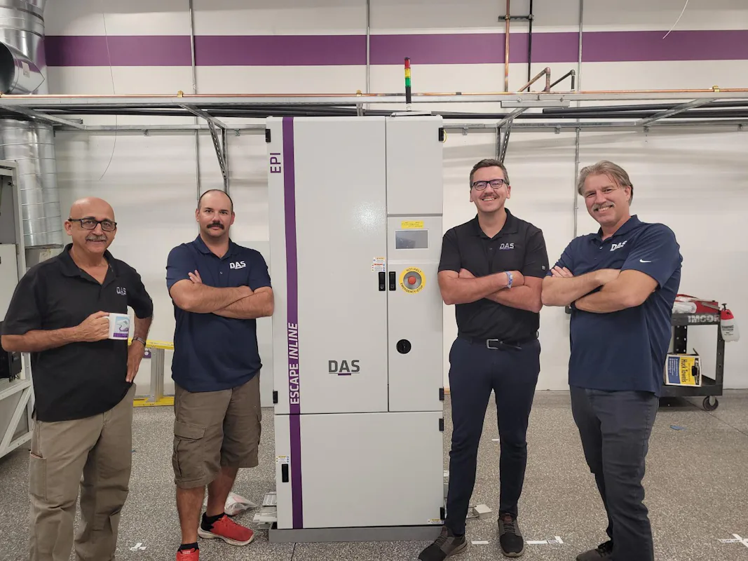 Mitarbeiter worldwide Four men in matching navy shirts stand confidently in front of a large industrial control panel, labeled "DAS" and "EPI," within a spacious room equipped with exposed wiring and ventilation pipes.
