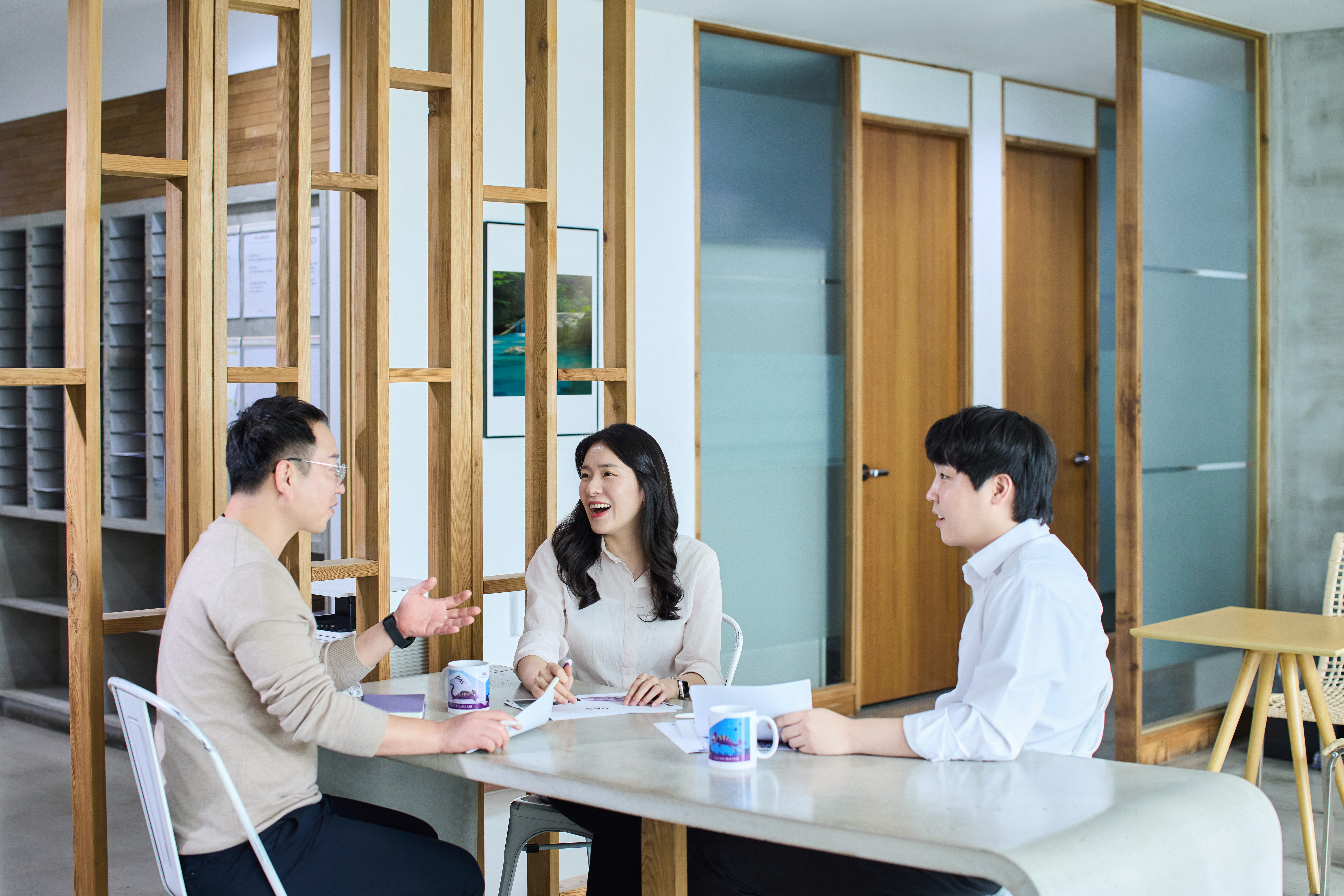 Three people are sitting at a modern office table, engaging in a discussion, with coffee mugs and documents on the table.