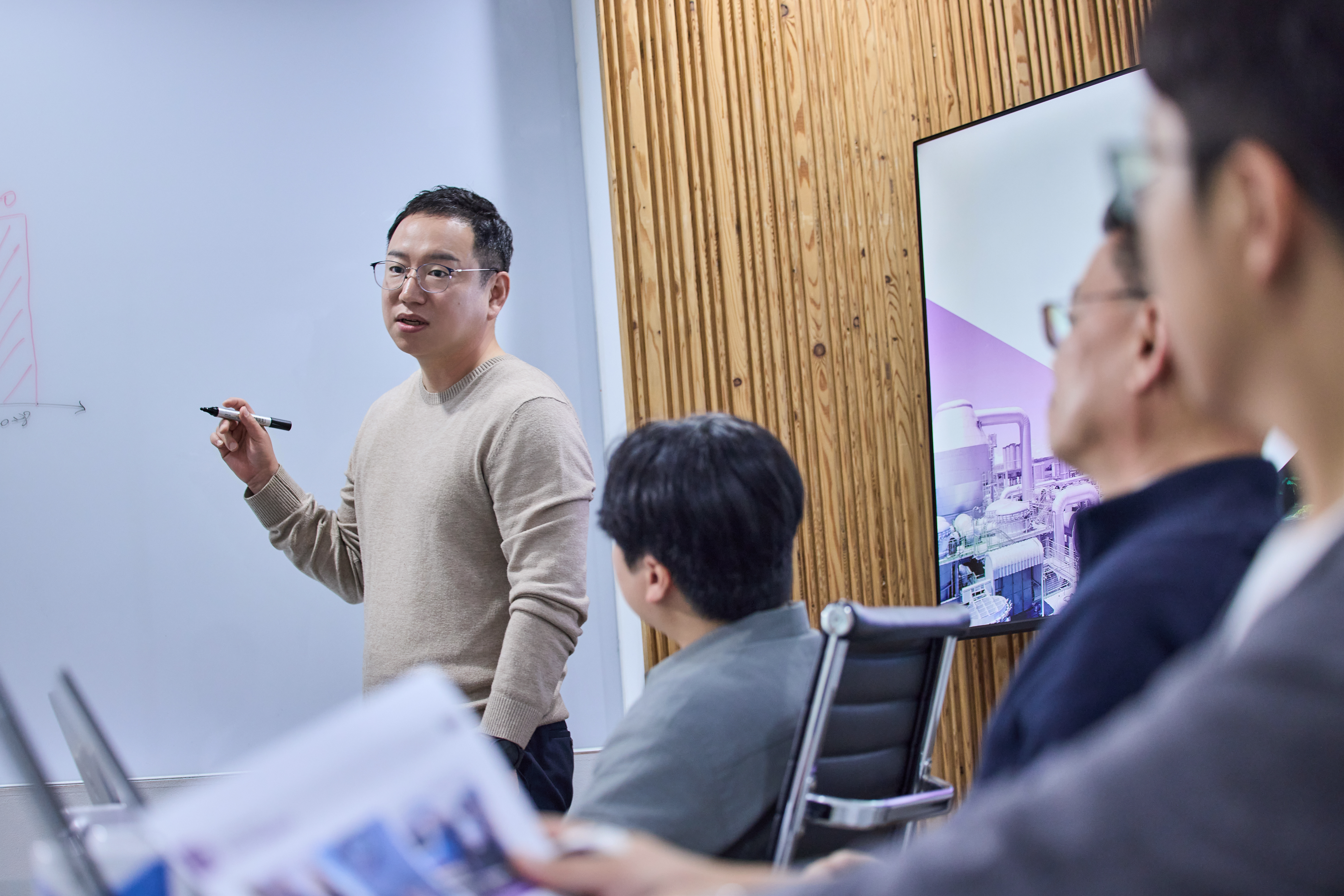 4 명의 직원이 현대적인 사무실의 회의 테이블에 둘러앉아 이야기를 나누고 있다. 가운데 있는 사람은 노트북을 들고 집중하고 있는 모습이다.