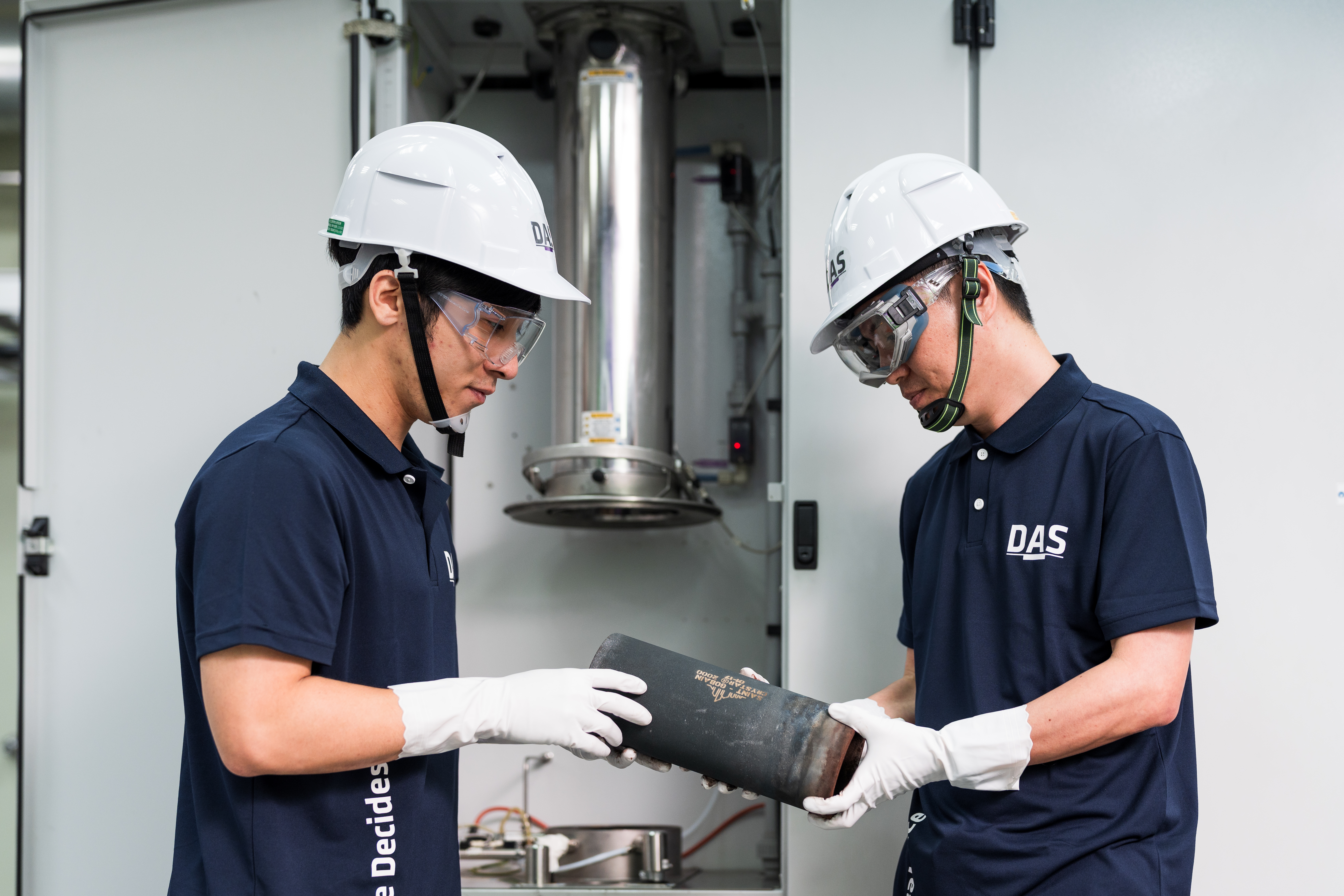 兩名穿著藍色制服和白色安全帽的工人在工業機械設備前協作檢查一個黑色圓柱型金屬部件，專注於其細節。