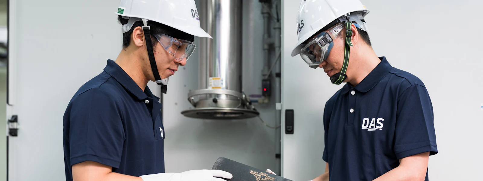 ISC 協作檢查 兩名穿著藍色制服和白色安全帽的工人在工業機械設備前協作檢查一個黑色圓柱型金屬部件,專注於其細節。