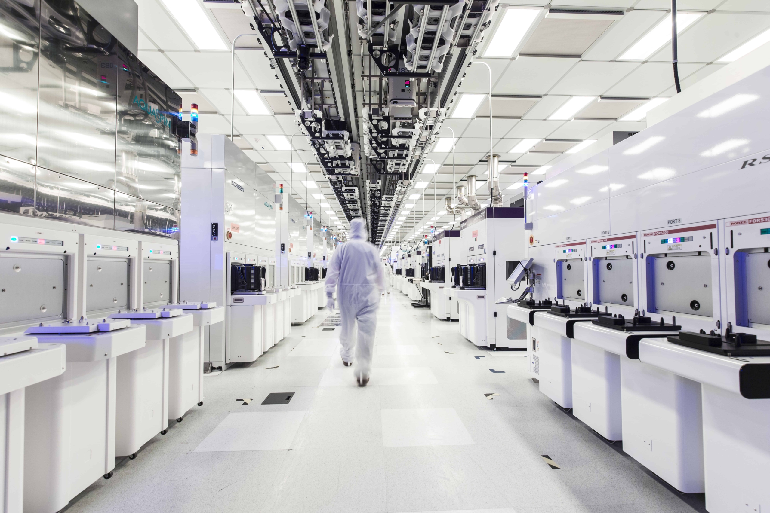 A technician in a white protective suit walks through a brightly lit clean room full of modern machines in a semiconductor factory.