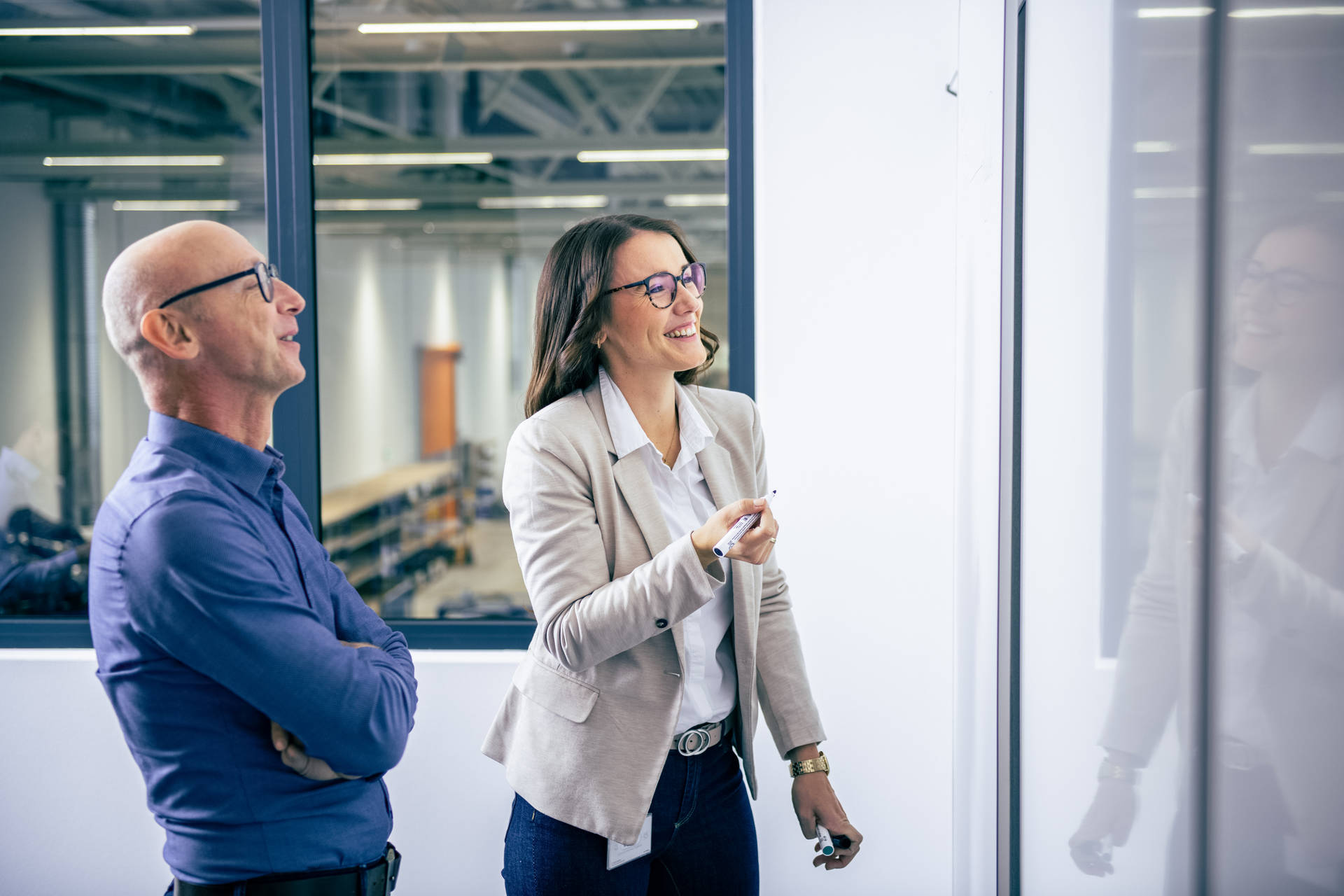 A man and a woman in a modern office stand in front of a whiteboard while the woman writes something on it and both smile.