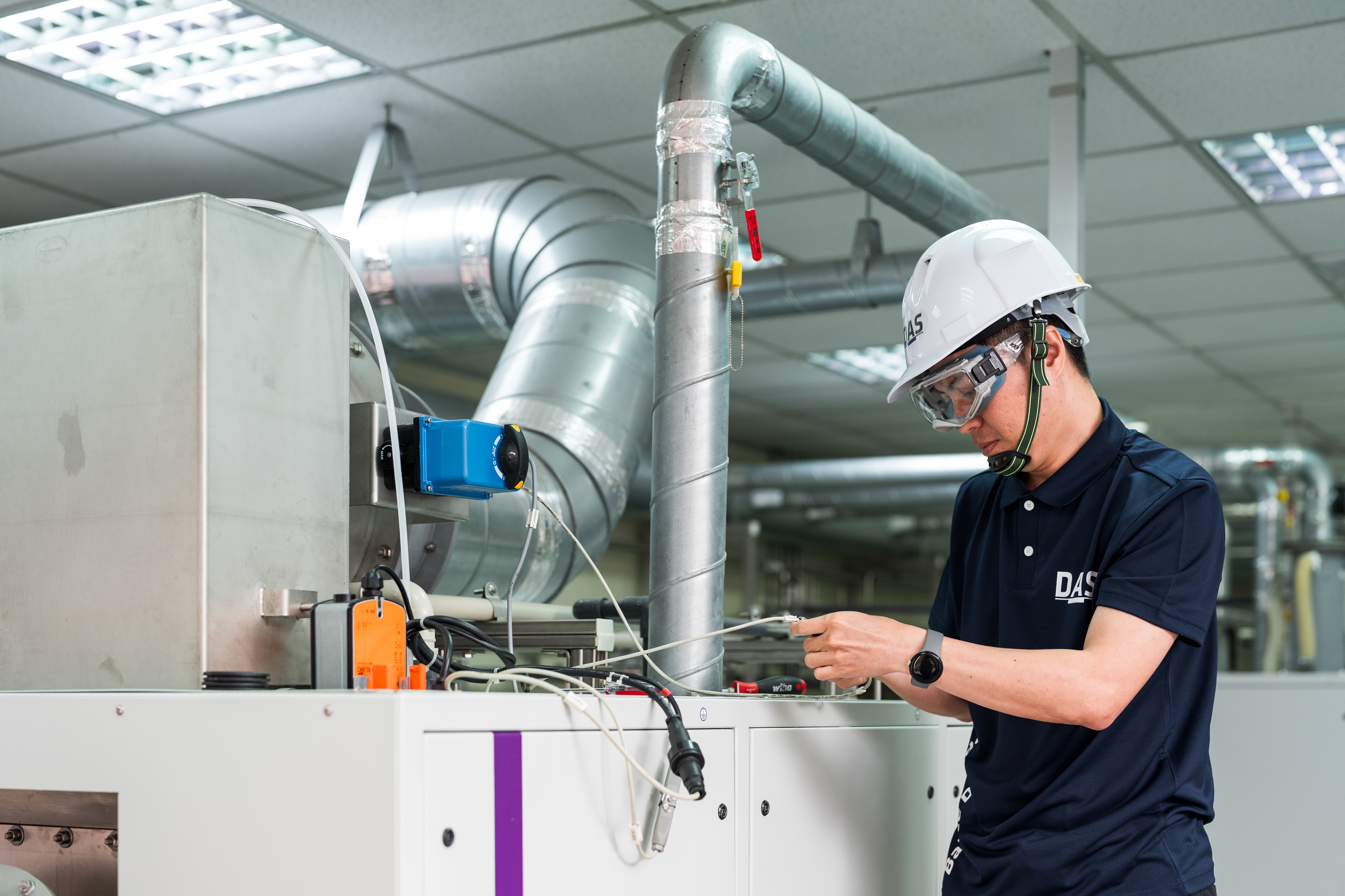 工廠中穿著工作服與安全帽的技術員正在檢查機械設備，周圍可見複雜的管道和儀表板，專注於機械保養工作。