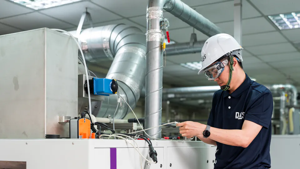 機台檢測 工廠中穿著工作服與安全帽的技術員正在檢查機械設備,周圍可見複雜的管道和儀表板,專注於機械保養工作。